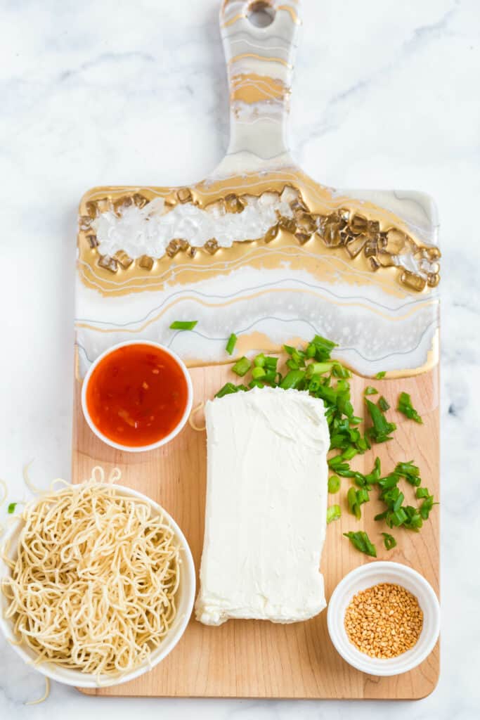 ingredients needed to make a thai chili cream cheese board: cream cheese, thai chili sauce, dry stir fry noodles, chopped green onions, toasted sesame seeds.