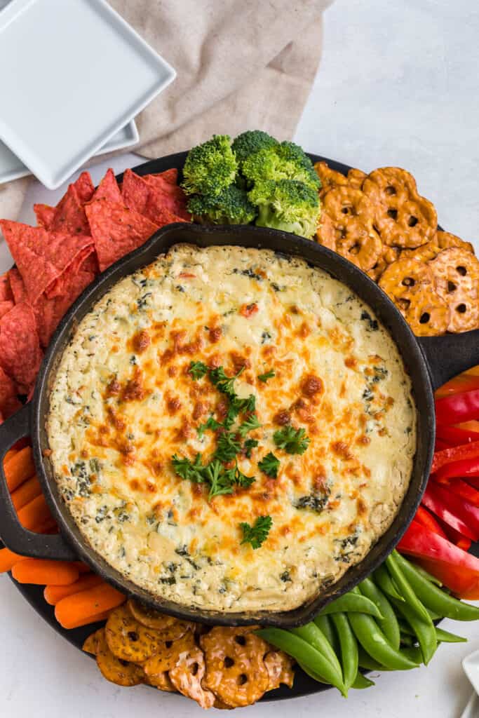 cheesy baked spinach dip in a cast iron skillet surrounded with colored tortilla chips, pretzel chips, and raw vegetables for dipping.