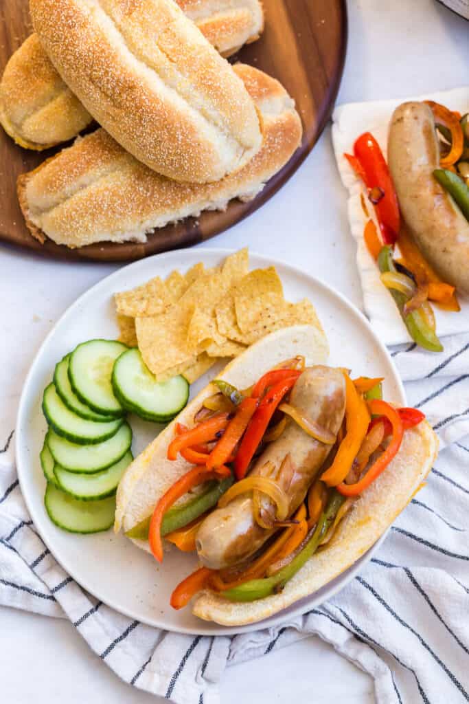 overhead view of cooked brats in a bun served with peppers, onions, cucumbers, and tortilla chips on the side.